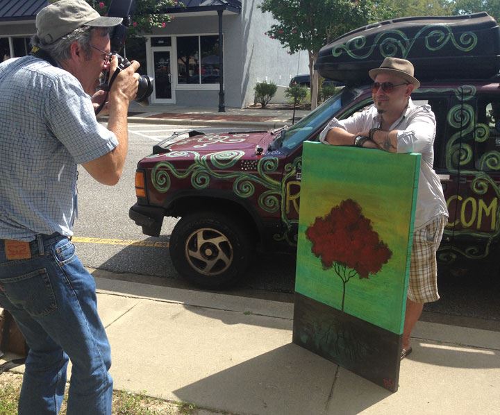 A photographer captures an artist posing with a painted canvas featuring a tree, in front of a colorful vehicle parked on the street.