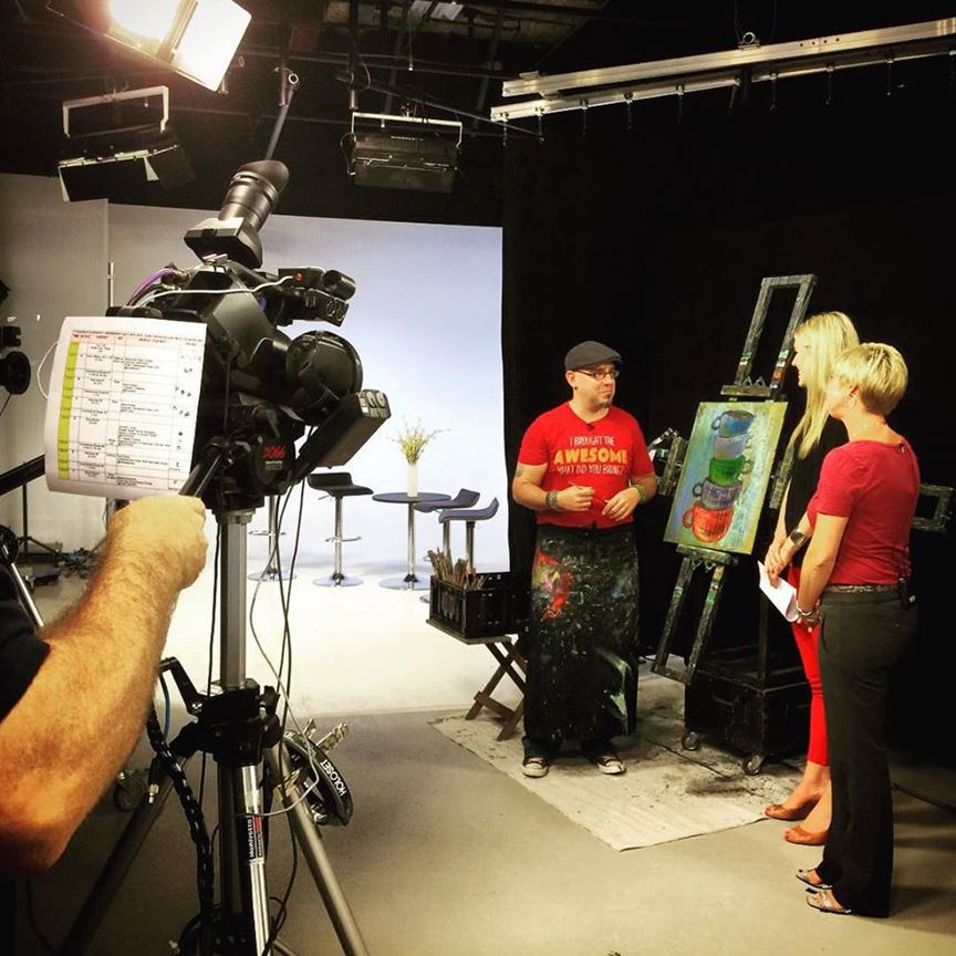 A television studio setting featuring an artist in a red shirt explaining his painting to two hosts. The artist stands beside an easel with a colorful artwork. A camera is focused on the scene, with lighting and studio equipment visible in the background.