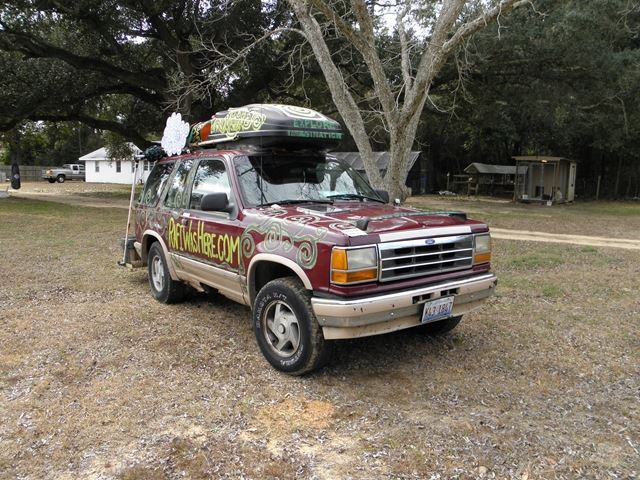 An old Ford SUV parked on a grassy area, featuring colorful decorative graphics on the body and a roof rack with items on top.