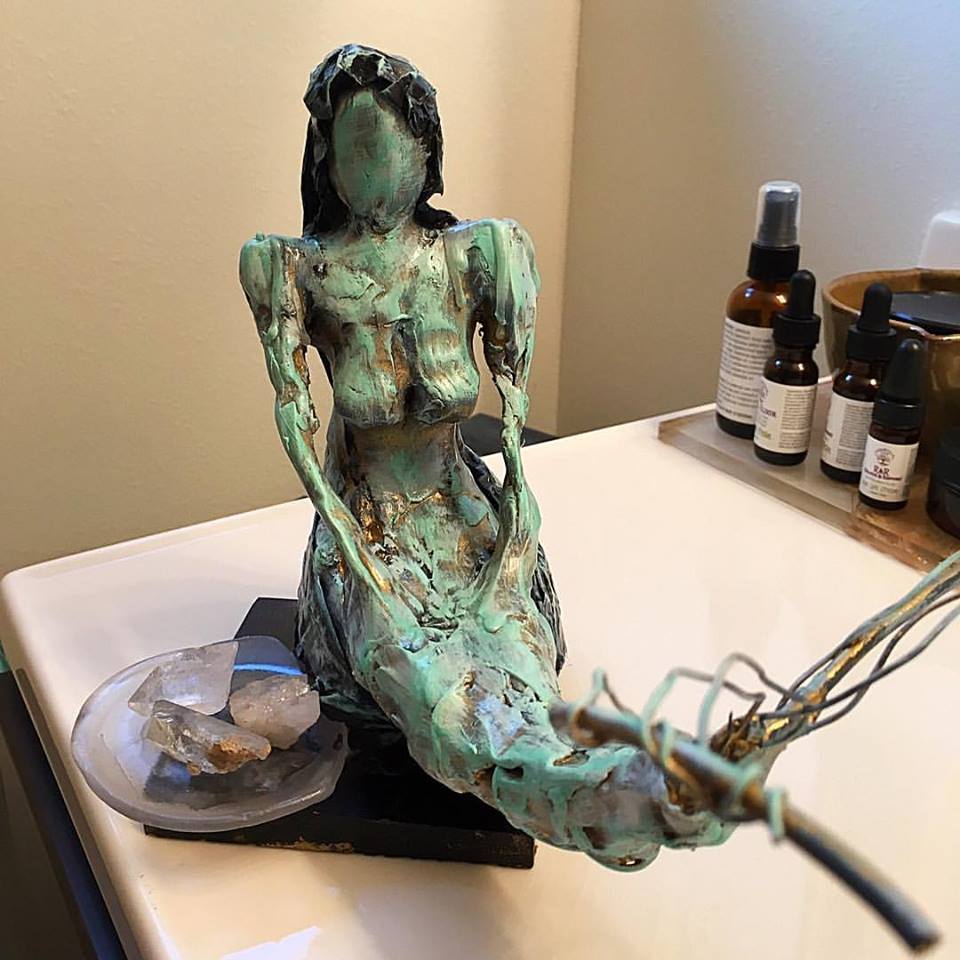 A detailed sculpture of a figure with a turquoise patina, positioned on a flat surface next to some crystal pieces and various bottles in the background.