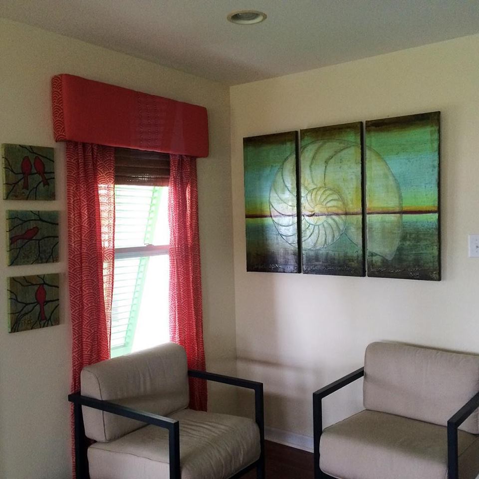 Cozy corner of a room featuring two beige chairs with metal frames, a large abstract painting of a nautilus shell mounted on the wall, and colorful red curtains framing the window.