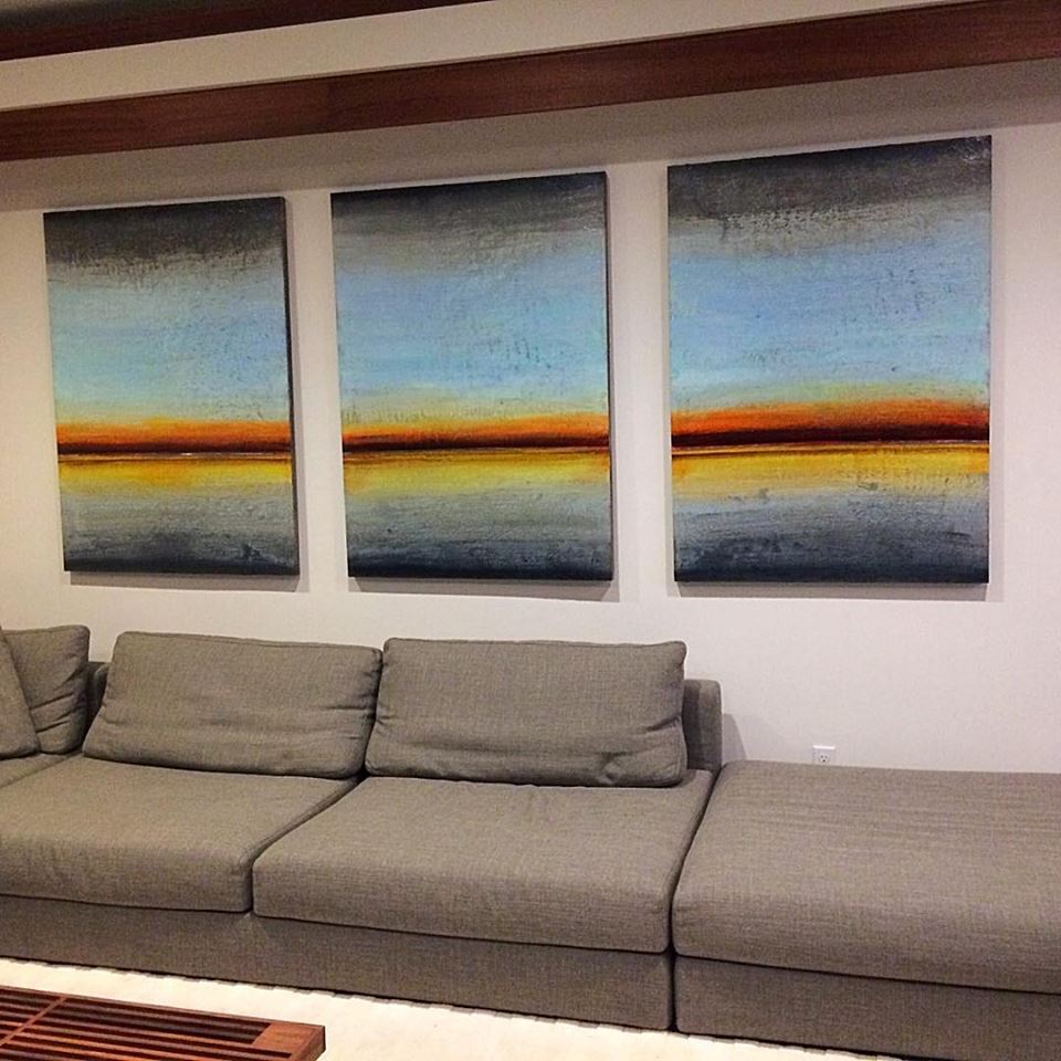 Three abstract landscape paintings with blue and orange tones hanging above a gray sectional sofa in a modern living room.