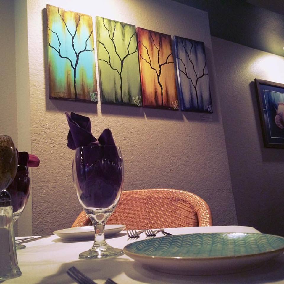 Table setting in a restaurant featuring a glass with a purple napkin, a green plate, and forks, with colorful tree paintings on the wall.