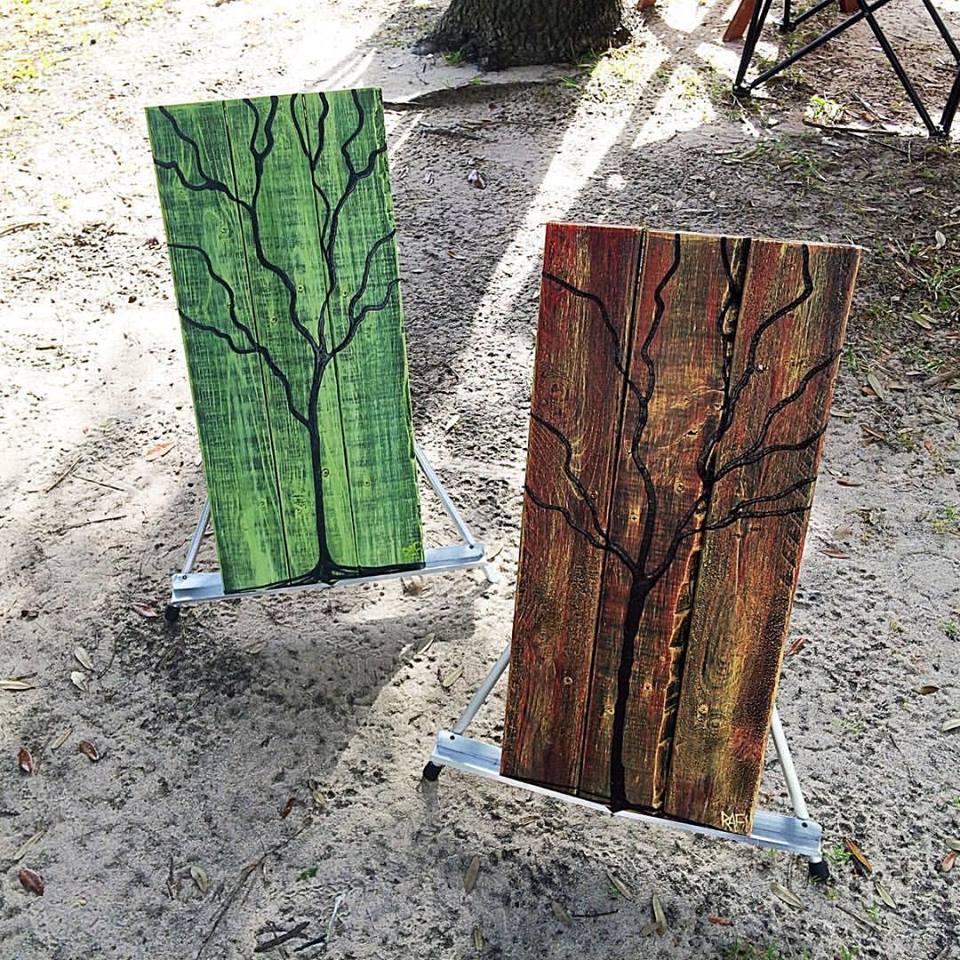 Two wooden paintings resembling trees, one in green with branch-like designs and the other in brownish-red with a similar pattern, displayed outdoors on easels in a sandy area.