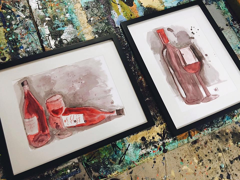 Two framed watercolor paintings of wine bottles and a glass set on a colorful, textured surface.