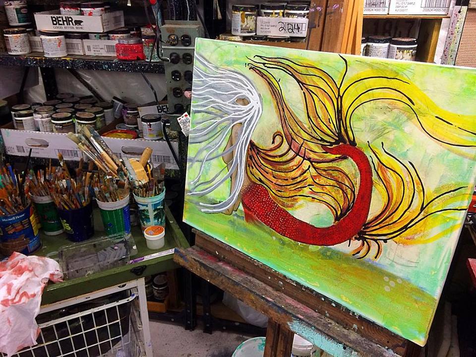 An abstract painting featuring vibrant colors with a prominent red fish-like form and flowing, yellowish strands against a green background, surrounded by art supplies and paint containers.