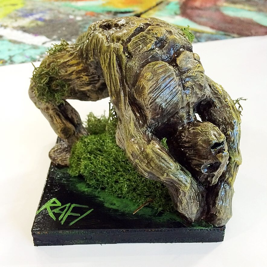 A detailed sculpture showing a humanoid figure merging with tree roots and moss, resting on a black base with the artist's name 'RAFI' painted in green.