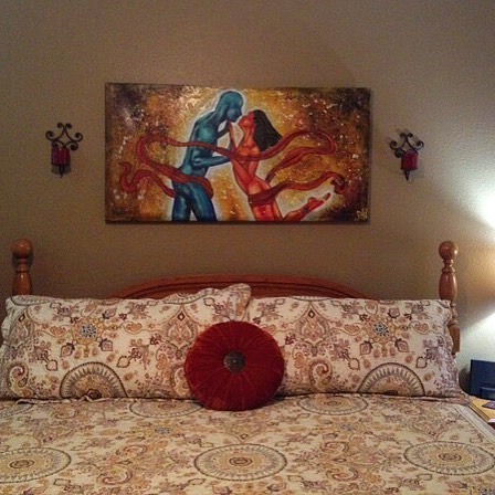 A cozy bedroom featuring a bed with decorative pillows and a patterned bedspread, adorned with a painting of a couple embracing, set against a warm-colored wall.
