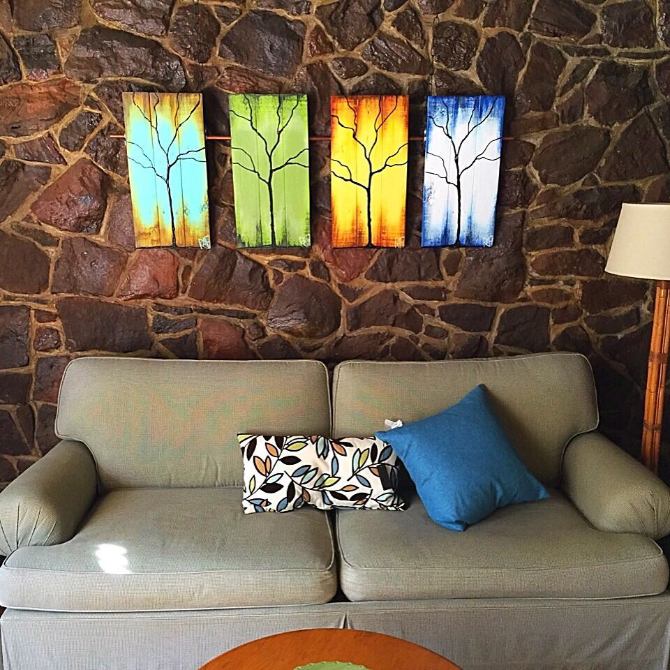 A cozy living room scene featuring a light sofa with decorative pillows, placed in front of a stone wall. Above the sofa are four vertical canvas paintings with tree designs in vibrant colors including blue, green, orange, and yellow.