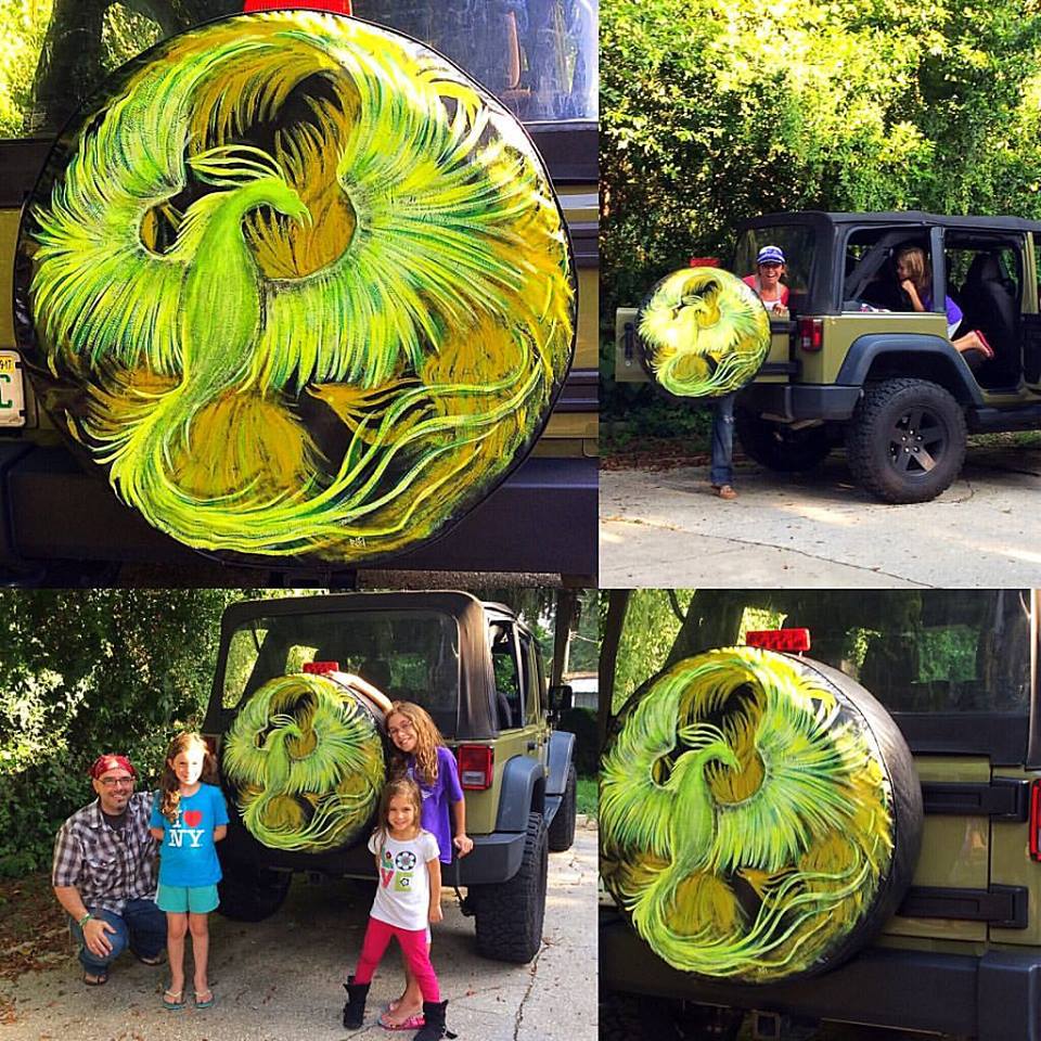 Colorful painted spare tire cover featuring a vibrant yellow and green phoenix design, displayed on the back of a Jeep. Children and an adult pose alongside the vehicle.