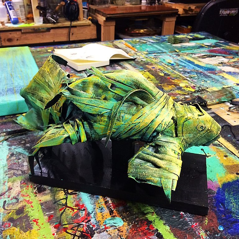Sculpture of a fish made from green fabric strips, displayed on a colorful messy workbench.