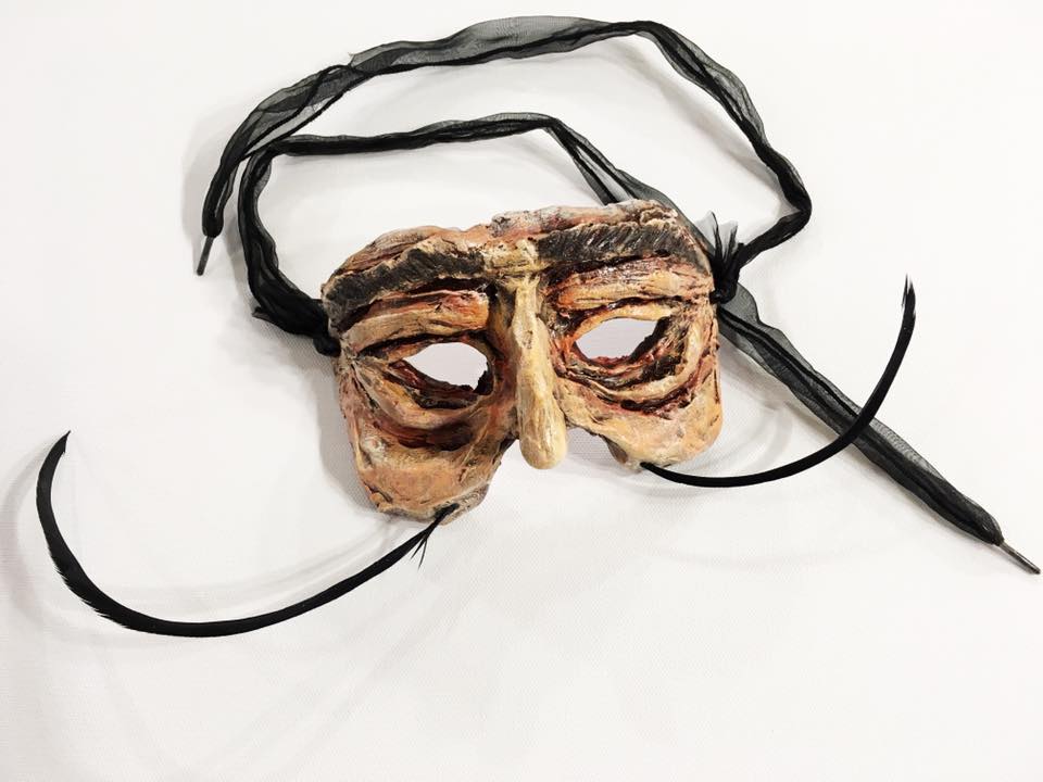A handcrafted mask featuring exaggerated facial features and a textured surface, adorned with black ribbons for securing it. The mask has an earthy color palette with hints of brown and a distinct artistic style.