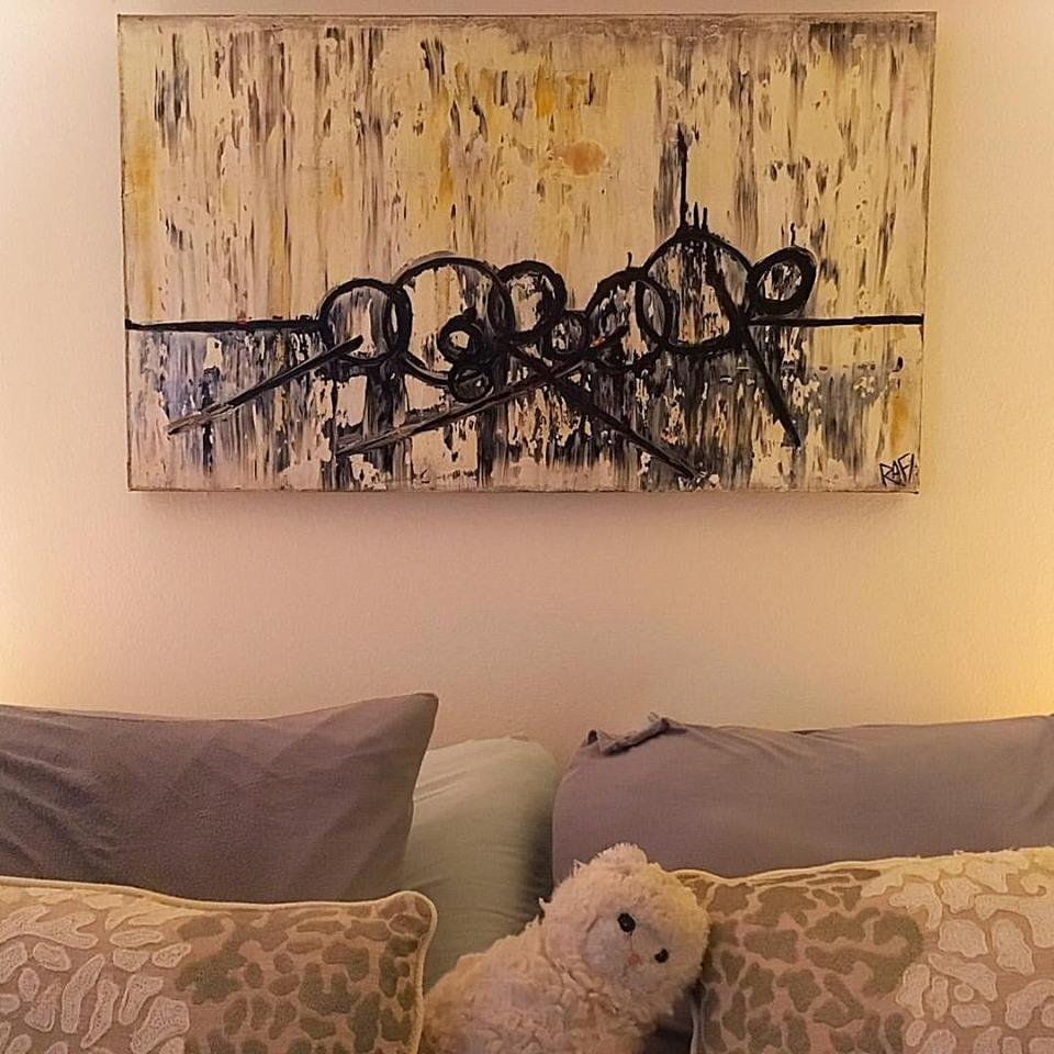 An abstract painting with circular and linear designs displayed above a bed with various pillows and a plush toy on the side.