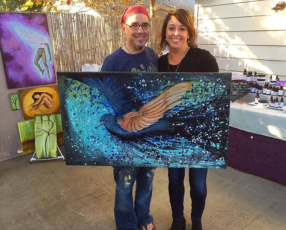 An artist posing with a woman, holding a vibrant painting of a bird in flight, with various artworks displayed in the background.