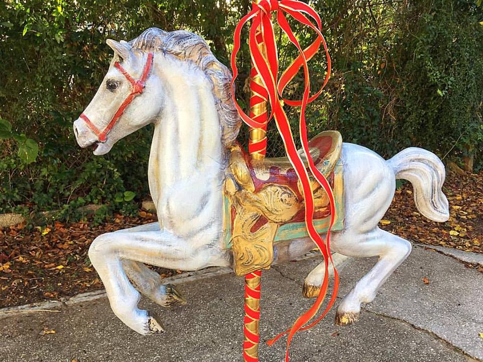A decorative carousel horse with a white body, red bridle, and colorful saddle, positioned against a backdrop of greenery.