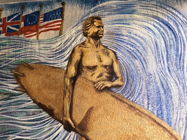 Mural depicting a muscular surfer holding a surfboard, with stylized waves and flags of the United Kingdom and the United States in the background.