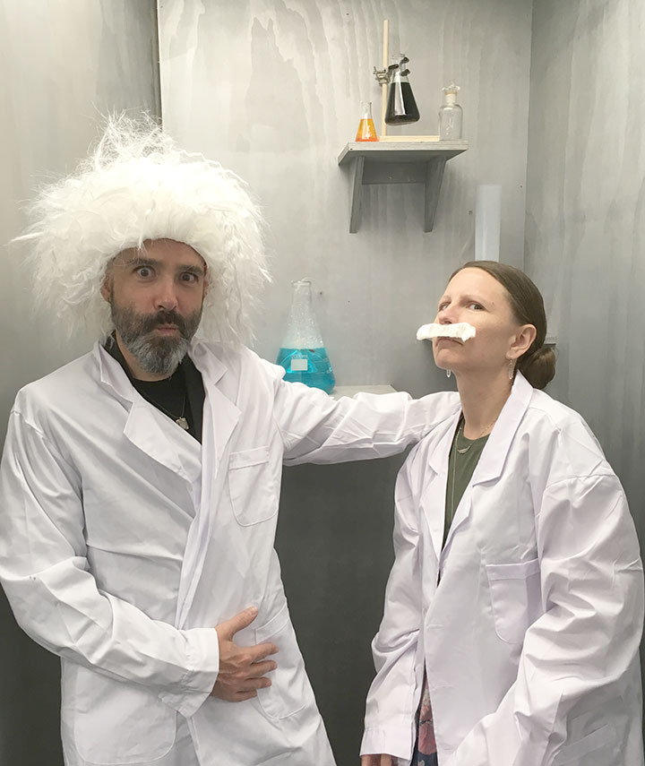 A man with a white wig and lab coat poses dramatically next to a woman in a lab coat wearing a fake mustache, with laboratory equipment in the background.
