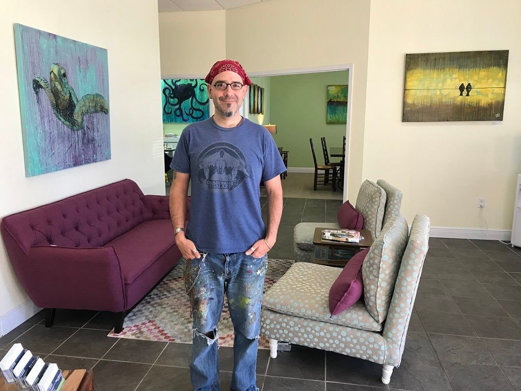 An artist stands in a brightly lit gallery space, wearing a bandana and a t-shirt. Colorful artwork is displayed on the walls, including a painting of a turtle. The artist has paint-splattered jeans and is positioned near a purple couch and patterned chairs.