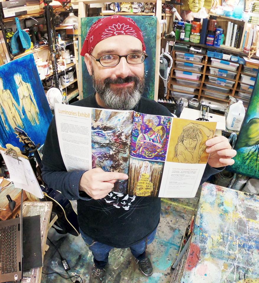 An artist with glasses and a red bandana smiles while holding an open magazine that displays various artworks, surrounded by a colorful studio filled with art supplies.