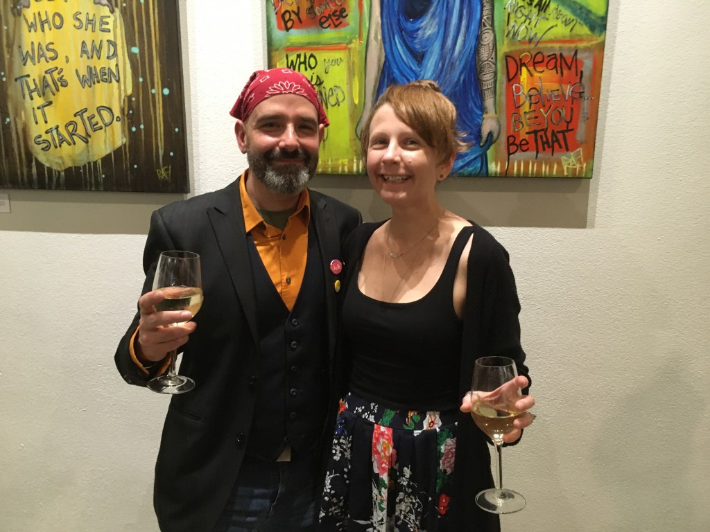 A man and woman posing together at an event, both holding glasses of wine. The man is wearing a black suit with an orange shirt and a red bandana, while the woman is in a black top and a floral skirt. Colorful artwork is visible in the background.