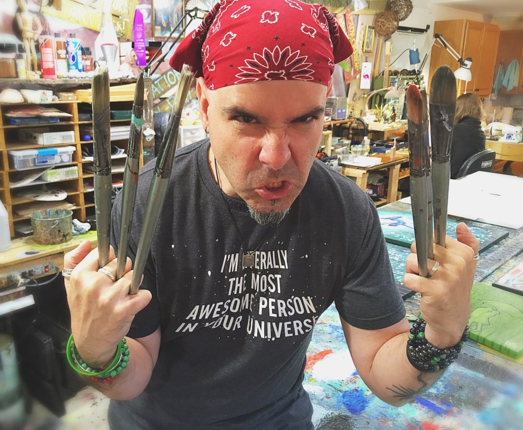 An artist in a workshop, wearing a red bandana and a t-shirt, displays paintbrushes in both hands while making a fierce expression.
