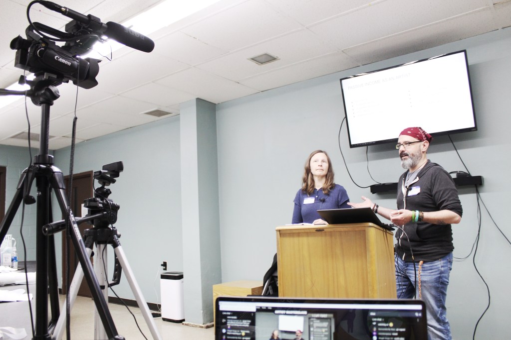 Two presenters standing at a podium in a classroom setting, with a projector screen displaying text behind them. A video camera is set up on a tripod in the foreground.
