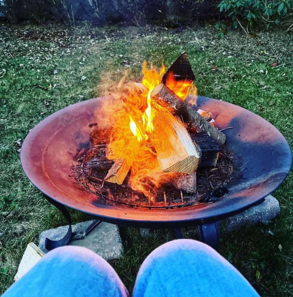 A close-up view of a fire pit with burning logs, surrounded by grass and a stone base, while a person's legs in jeans are visible in the foreground.