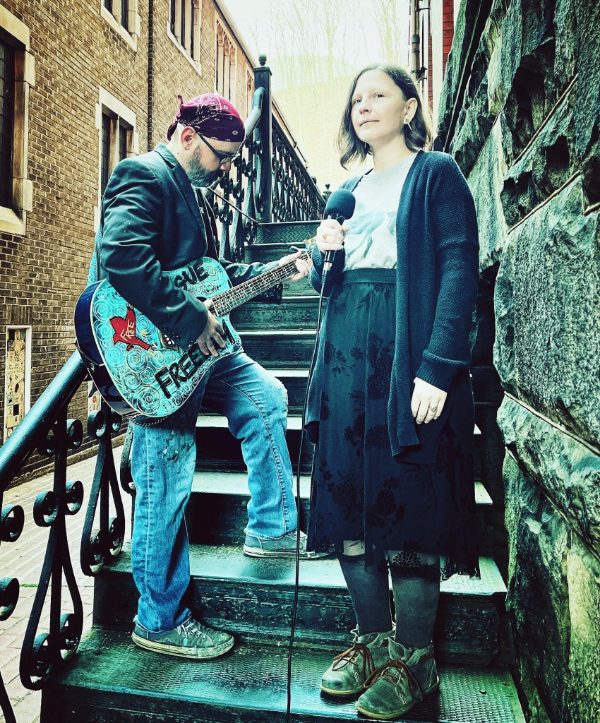 A man wearing sunglasses and a bandana plays a decorated guitar on a staircase, while a woman holding a microphone stands beside him, both in an outdoor setting with brick walls.