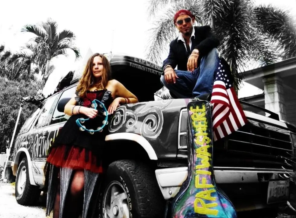 A man sitting on the hood of a decorated SUV, wearing a bandana and sunglasses, alongside a woman standing beside him, playing a tambourine. The scene is set in a tropical area with palm trees in the background, and an American flag is visible on the vehicle.