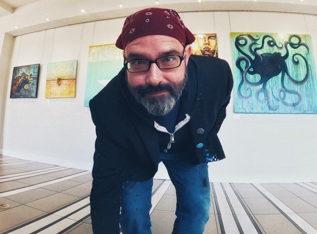 A man with a beard and glasses, wearing a bandana and a black blazer, poses in an art gallery filled with colorful paintings, including an octopus artwork in the background.
