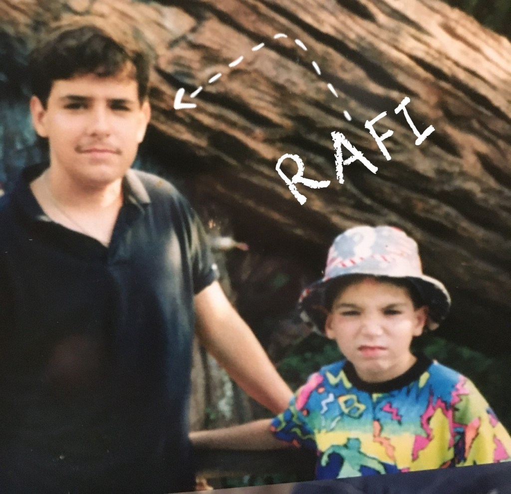 Two boys standing next to each other in front of a large tree. One boy is wearing a colorful shirt and a patterned hat, while the other is in a black polo shirt.
