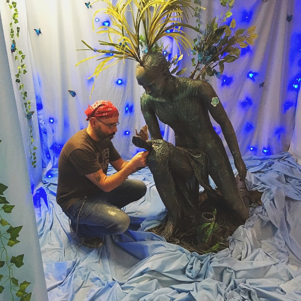 A person kneeling and painting a tree-like sculpture in a decorated space with blue background and soft lighting.