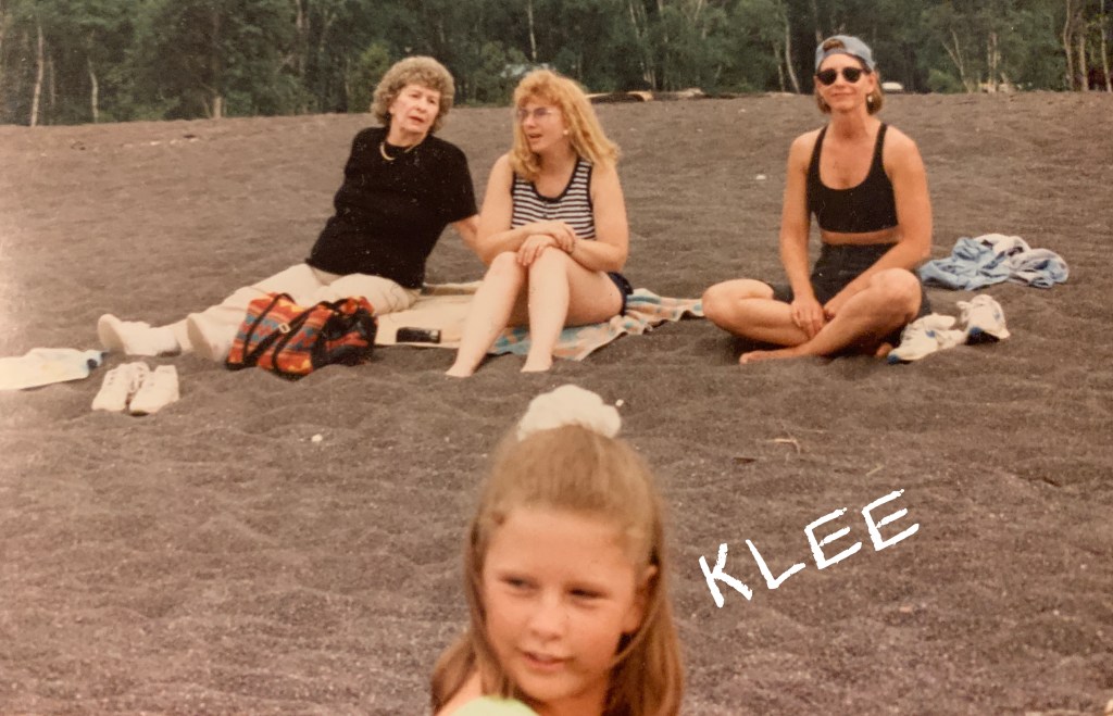 A group of three women sitting on a sandy beach, with one woman in the foreground focusing on the camera. The background features trees and a rocky surface, suggesting a beach setting.