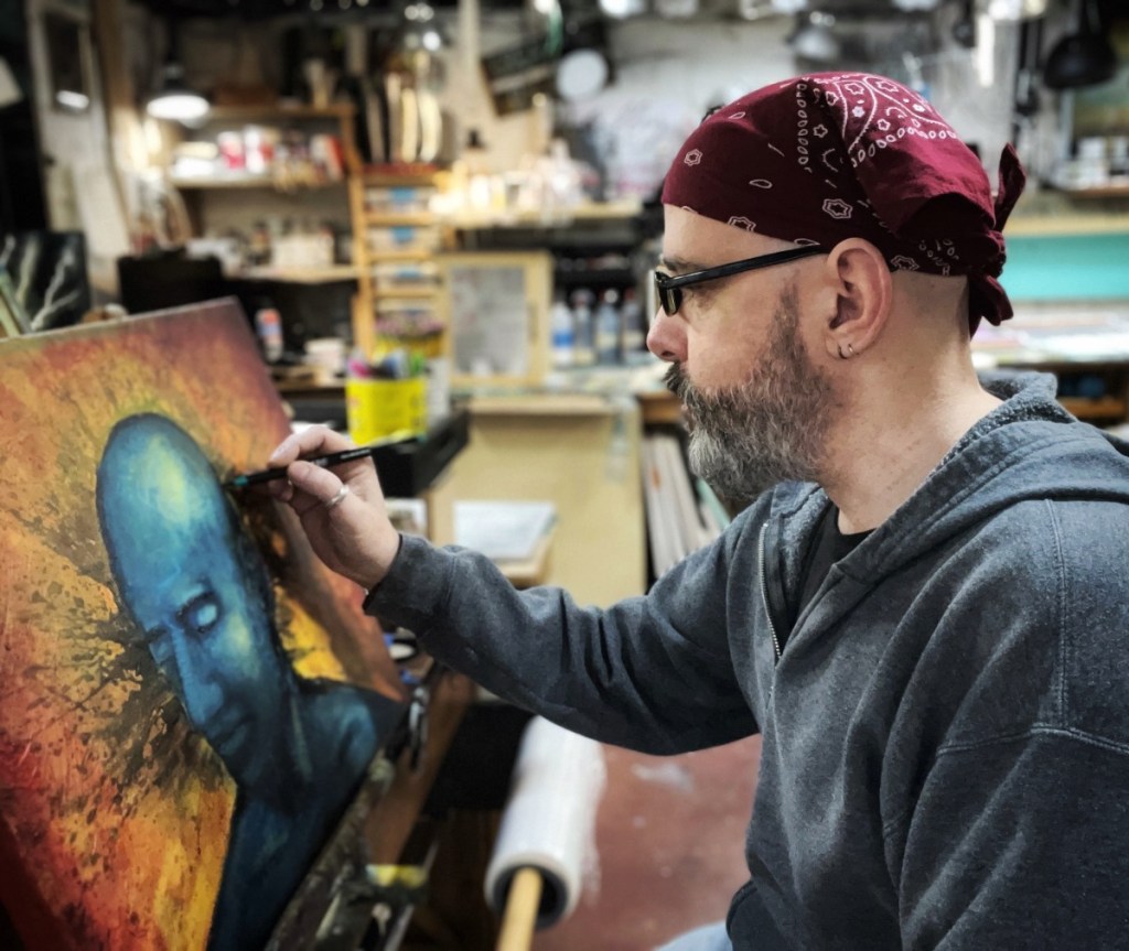A person painting a blue figure on canvas in an art studio, wearing a red bandana and glasses.