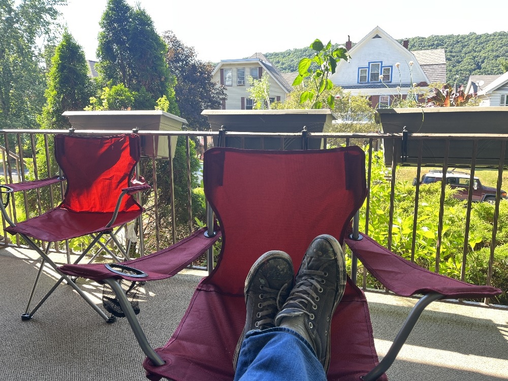 A person's feet resting on a balcony railing with two red folding chairs in the background and a view of houses and greenery.