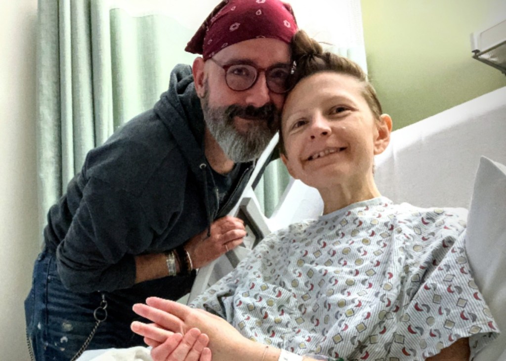 A man with glasses and a bandana stands next to a woman in a hospital gown, both smiling and looking at the camera.