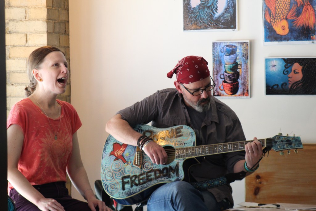 A woman singing passionately while a man plays an acoustic guitar next to her. The man is wearing a red bandana and glasses, and the guitar is decorated with the word 'FREEDOM.' Colorful artwork hangs on the wall behind them.