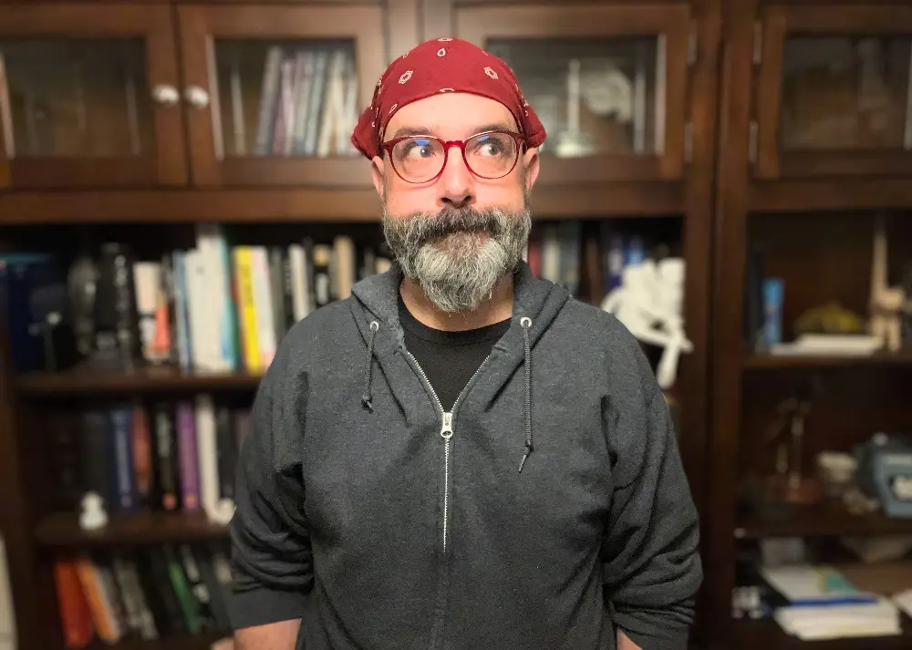 A person with glasses and a bandana, wearing a gray hoodie, standing in front of a bookshelf filled with various books and objects, displaying a thoughtful expression.