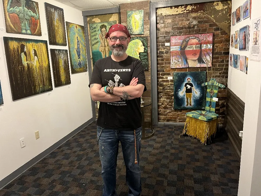 An artist standing in a vibrant gallery filled with colorful paintings and unique artwork, including a decorated chair and an exposed brick wall.