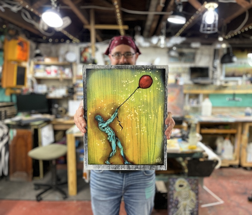 A person holding an artwork featuring a blue figure reaching for a red balloon against a green and orange background, inside an art studio.