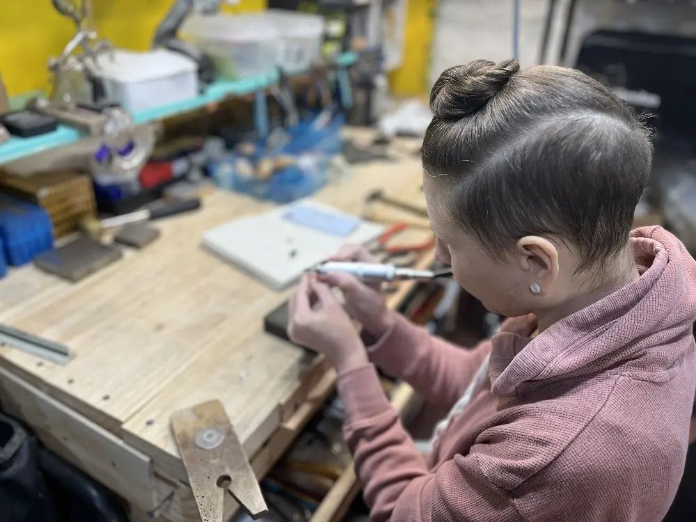 A person with short hair and a pink hoodie concentrating on a creative project at a workbench filled with tools and materials.
