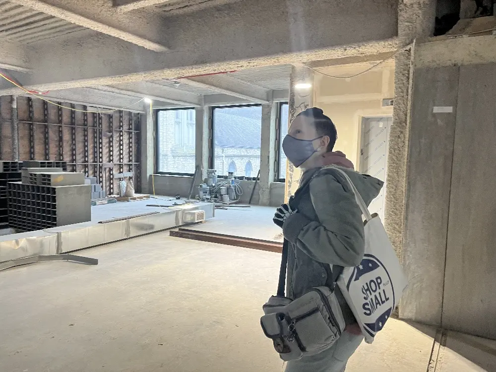 A person wearing a mask and gloves stands in a partially constructed space, holding a bag labeled 'SHOP SMALL'. The environment features exposed walls, windows, and construction materials.