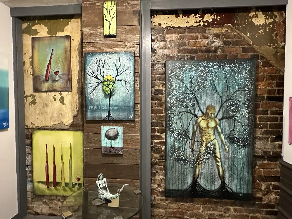 A cozy studio wall featuring various artworks, including paintings of trees, bottles, and unique figures, set against rustic brick and wooden backgrounds.