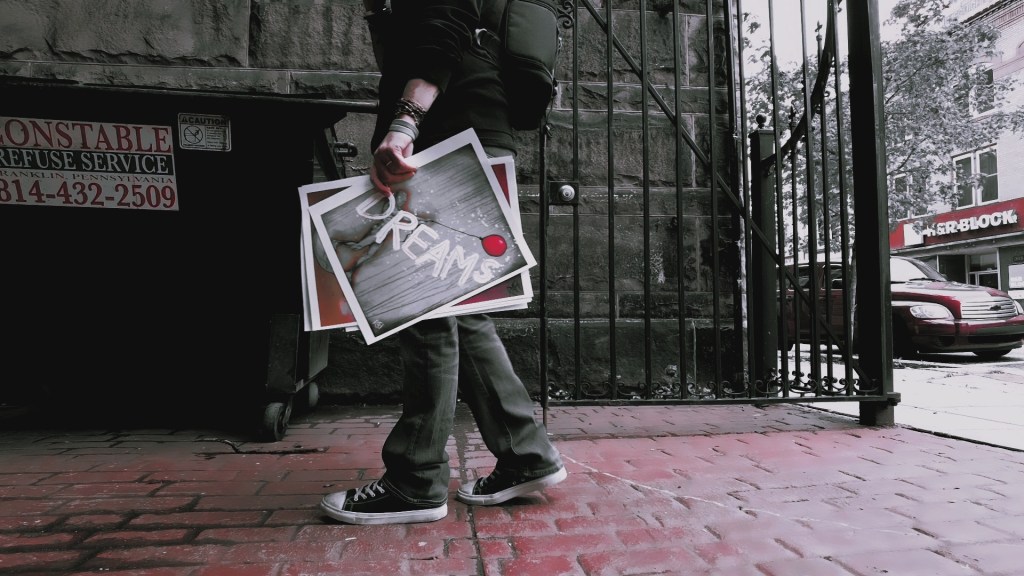 A person walking with photographs in hand, one of which prominently displays the word 'DREAMS', in an urban setting with brick pavement and a dumpster nearby.