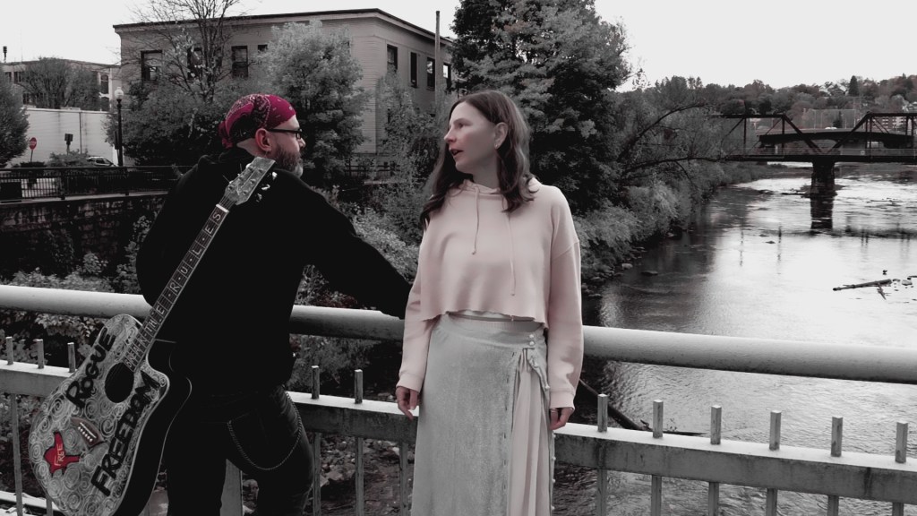 Two musicians standing by a river in Oil City, one wearing a black jacket and a bandana with a guitar, while the other is in a light-colored outfit, both engaged in conversation.