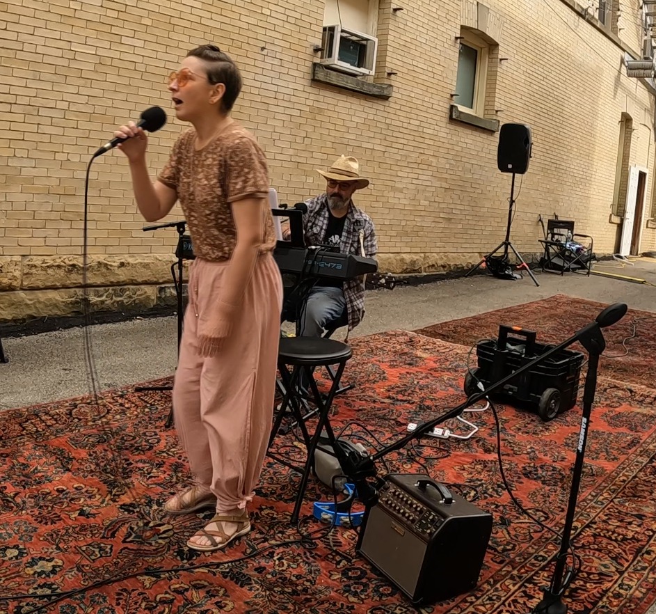 A female singer passionately performs into a microphone while wearing a brown patterned shirt and pink pants, with a male musician playing keyboard in the background. The performance takes place outdoors on a decorative rug against a brick wall.