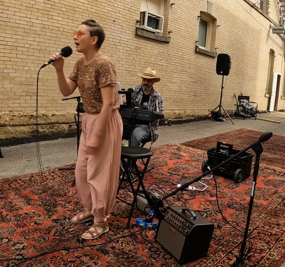 A female singer passionately performs into a microphone while wearing a brown patterned shirt and pink pants, with a male musician playing keyboard in the background. The performance takes place outdoors on a decorative rug against a brick wall.