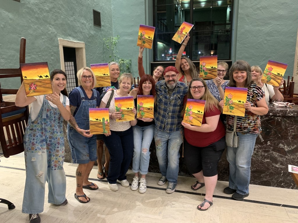 A group of happy individuals posing together, each holding their own painted canvas featuring a desert landscape with flowers and camels, showcasing a fun and creative art class experience.