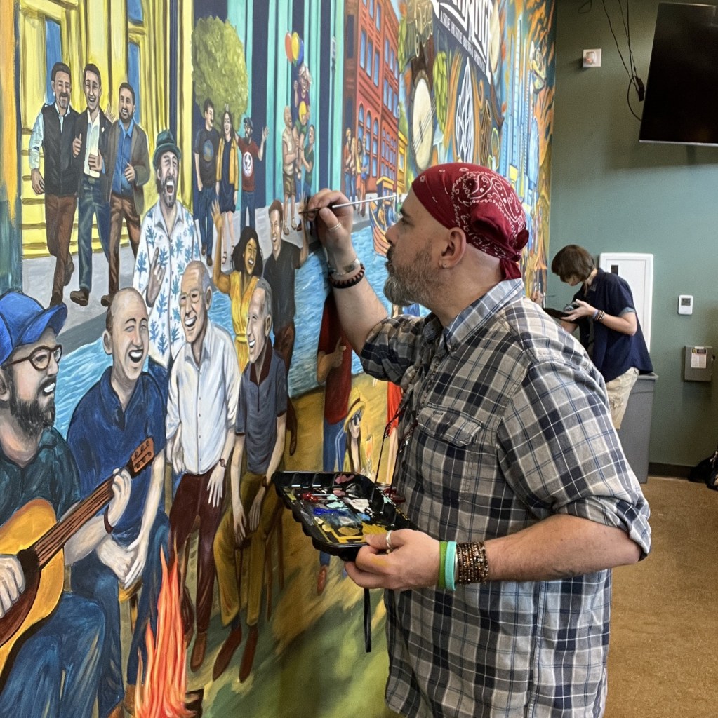 An artist with a bandana is painting a colorful mural featuring diverse people enjoying music and socializing. The scene captures a lively atmosphere with laughter and creativity.