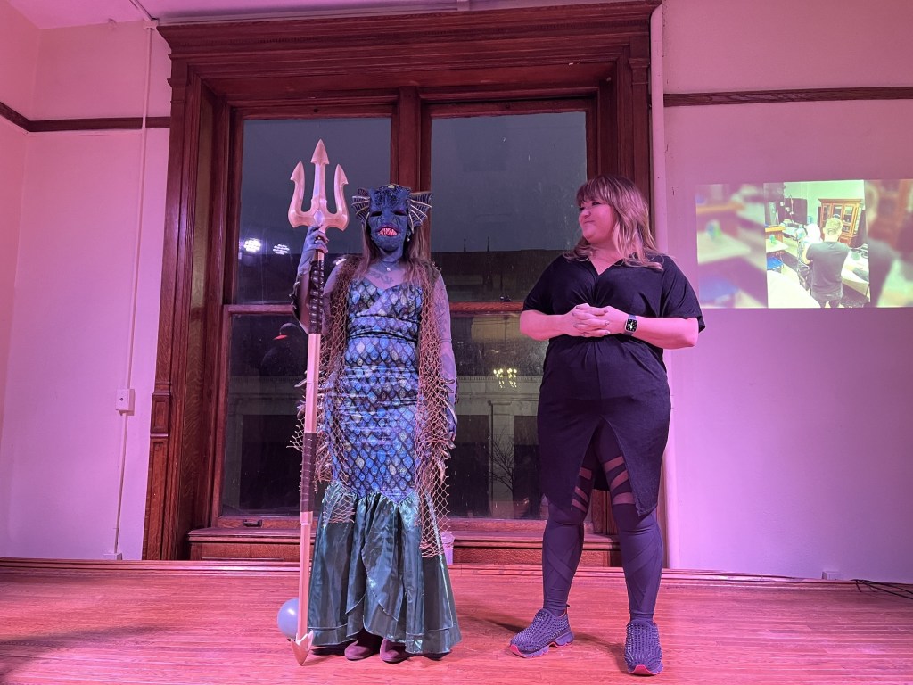 A performer dressed as a sea creature with scales and a trident stands on stage next to a woman in dark clothing, both engaged in a special effects makeup competition.
